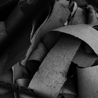 Strips of rubber, dirty, industrial and abstract looking, in a dark black and white photo, with the word "never" inscribed in a stretched, black font over a black wire that crosses the bottom left corner of the image.